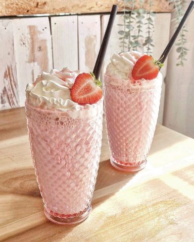 Two tall glasses of strawberry milkshake topped with whipped cream and a strawberry.